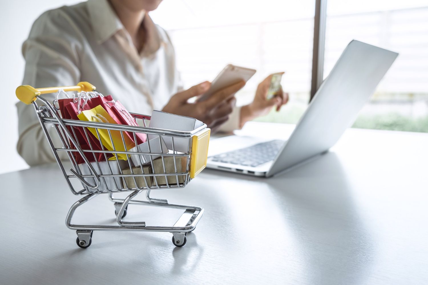 Woman holding phone and credit card and shopping online with a little shopping cart in foreground